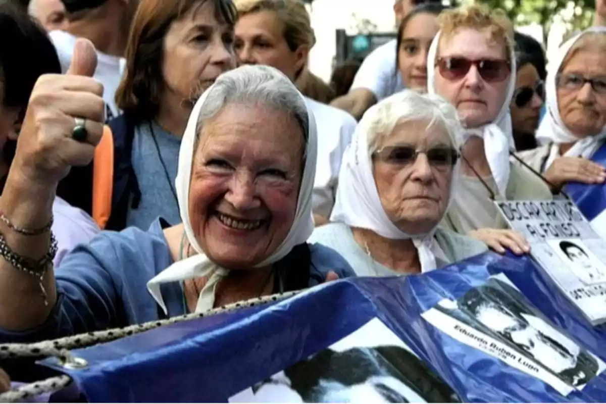 Un grupo de mujeres mayores con pañuelos blancos en la cabeza participa en una manifestación sosteniendo pancartas con fotografías.