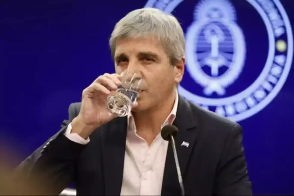 Un hombre de cabello canoso bebe agua de un vaso frente a un micrófono con un fondo azul que muestra un emblema oficial.