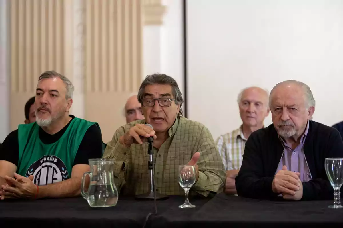 Un grupo de hombres sentados en una mesa durante una conferencia de prensa, uno de ellos hablando por un micrófono.