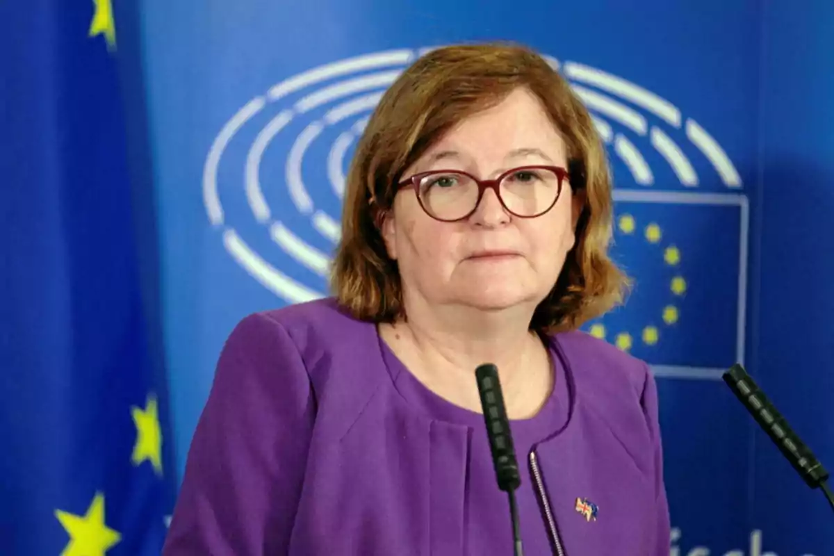 Mujer con gafas y chaqueta morada hablando frente a micrófonos con un fondo del Parlamento Europeo.