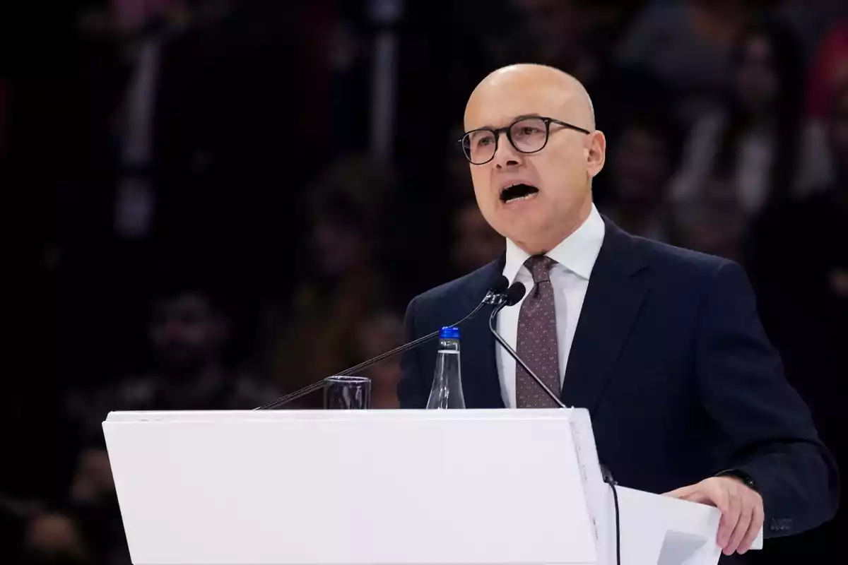 Un hombre con gafas y traje oscuro habla en un podio blanco durante un evento.