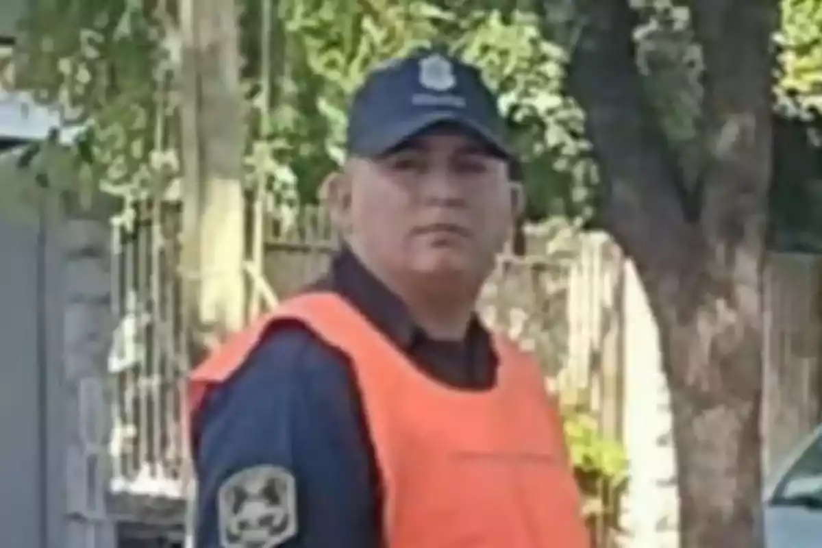 Un hombre con chaleco naranja y gorra está de pie al aire libre cerca de un árbol.