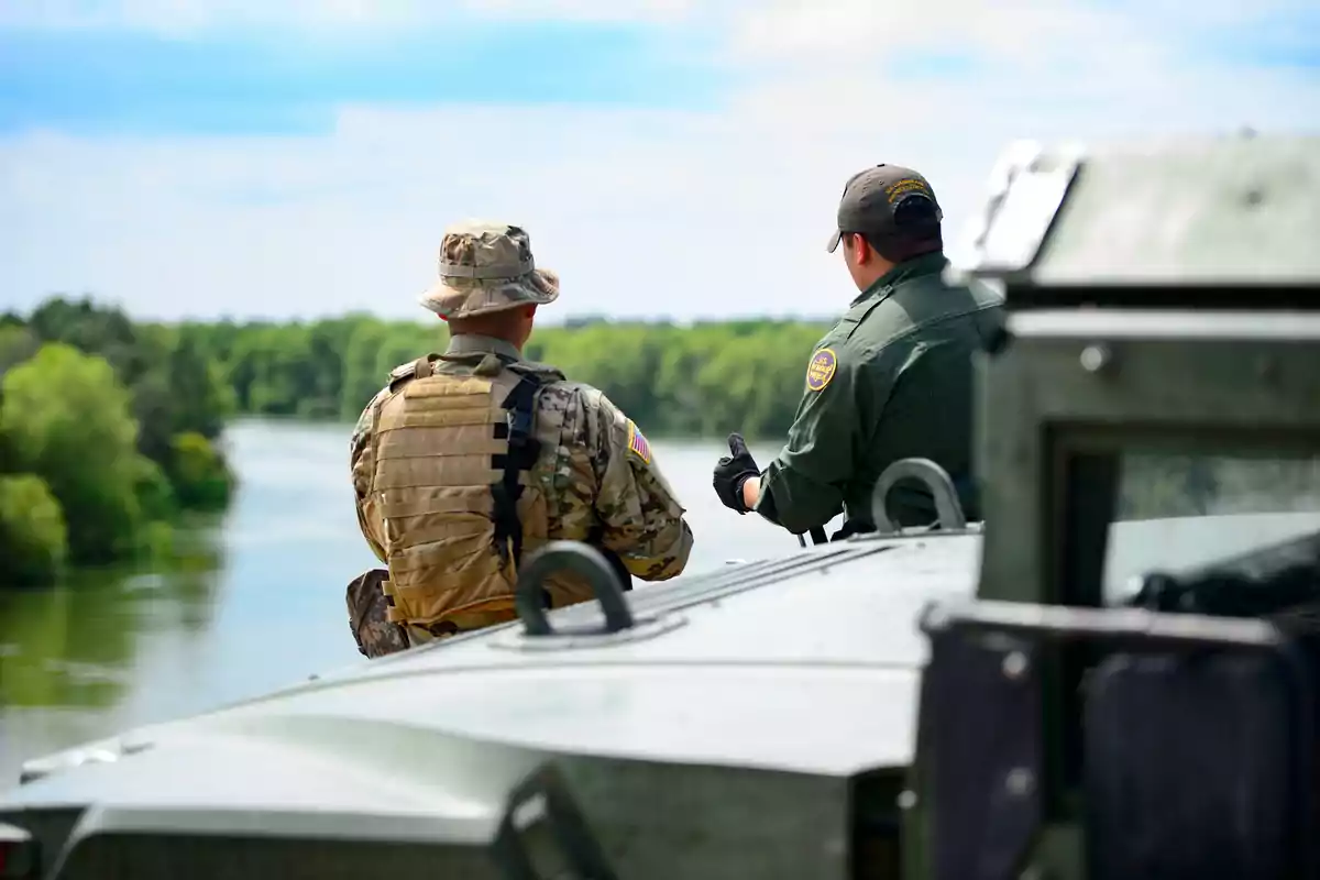 Dos personas con uniformes militares observan un río desde un vehículo blindado en un entorno natural.