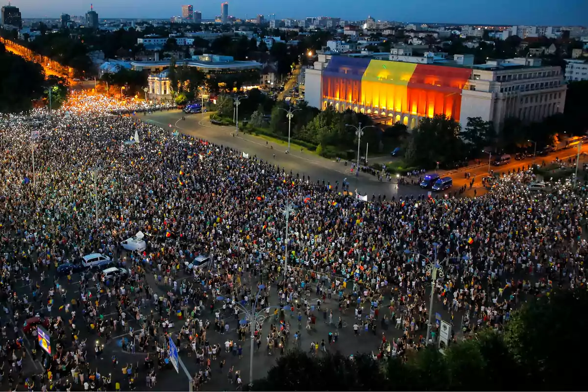 Una multitud se reúne en una plaza frente a un edificio iluminado con los colores del arcoíris durante el atardecer.