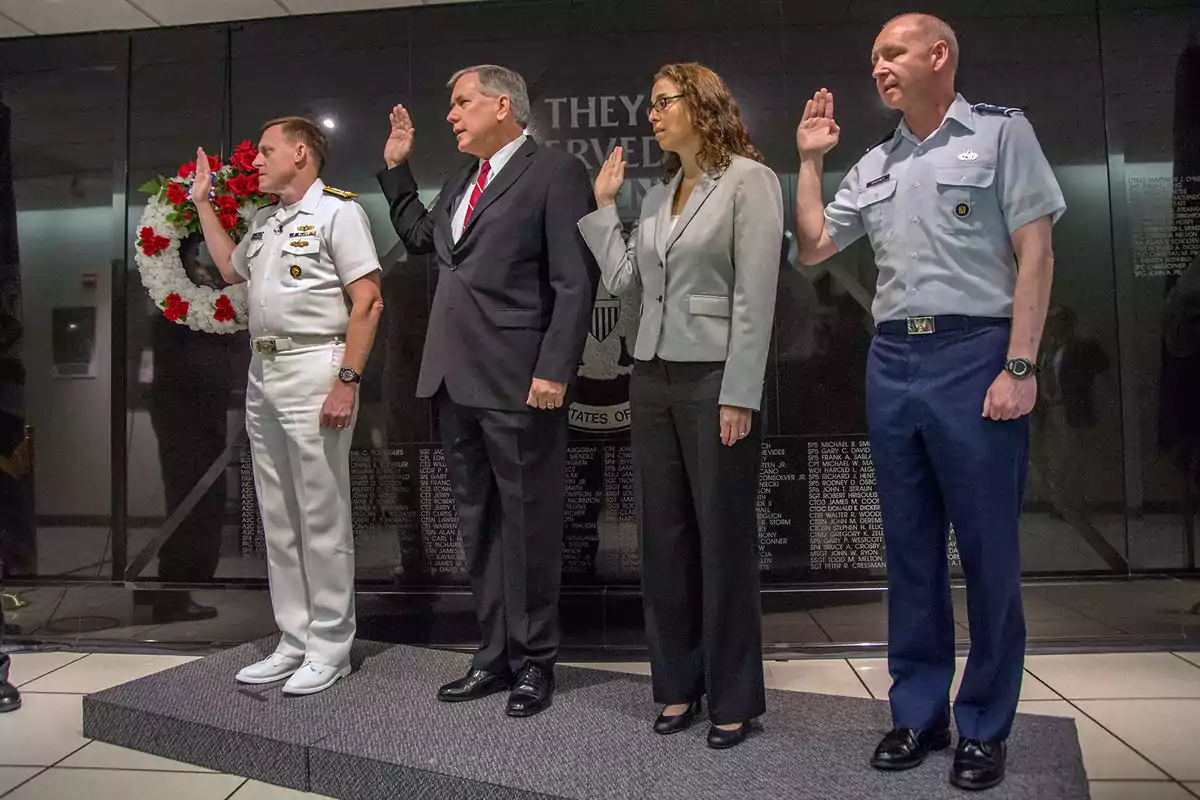 Cuatro personas de pie con la mano derecha levantada en un gesto de juramento, dos de ellas en uniforme militar y dos en traje formal, frente a un fondo oscuro con una corona de flores.