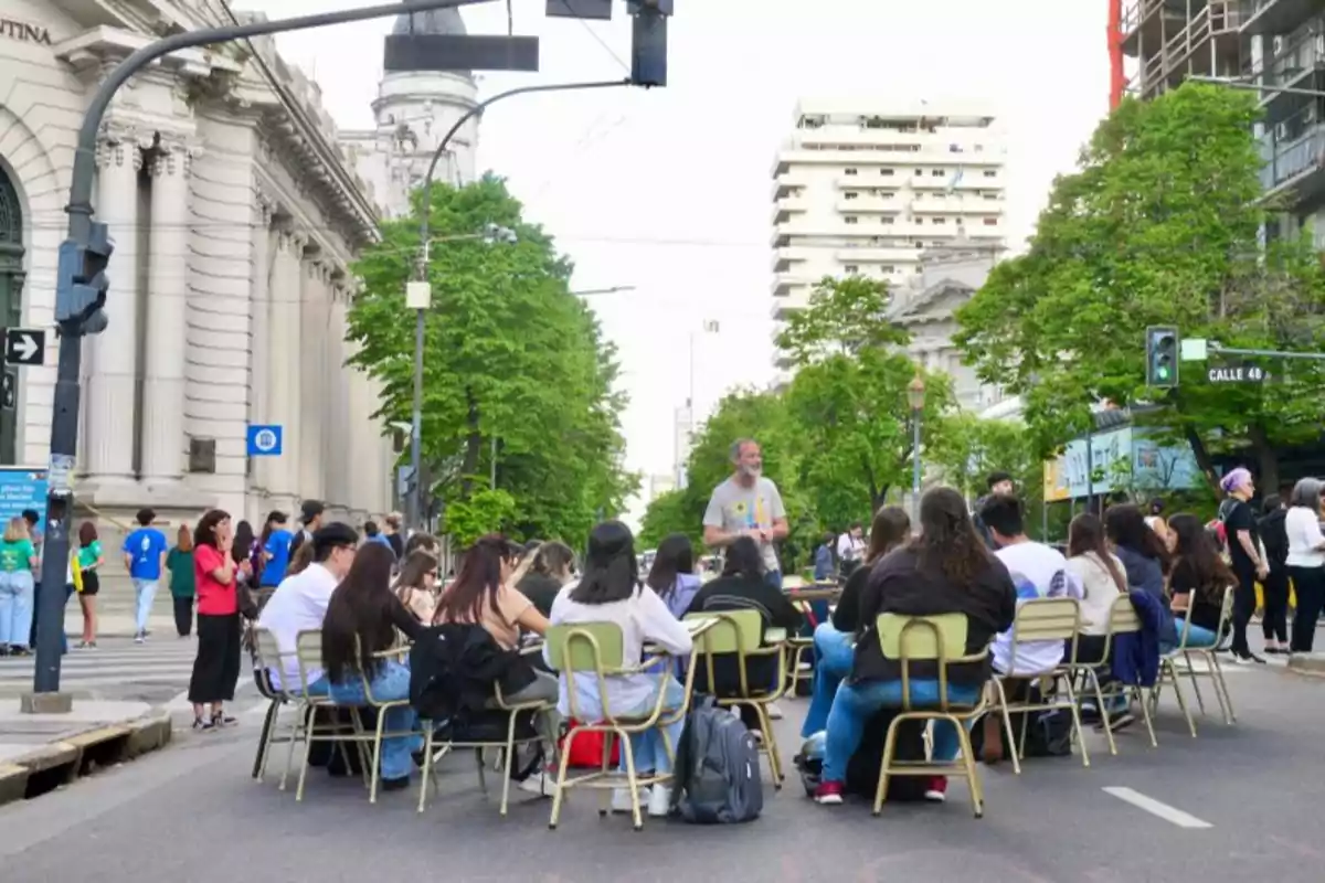 Un grupo de personas sentadas en sillas y escritorios en medio de una calle, con edificios y árboles alrededor.