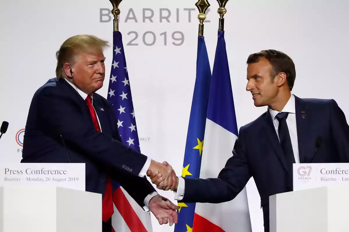 Two people shaking hands at a press conference with flags in the background.