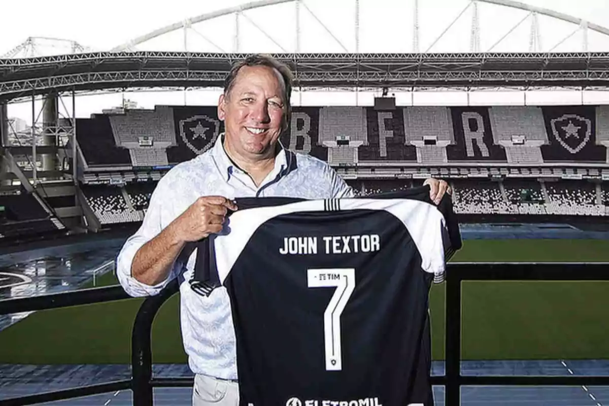 Hombre sonriendo mientras sostiene una camiseta de fútbol con el nombre "John Textor" y el número 7 en un estadio.