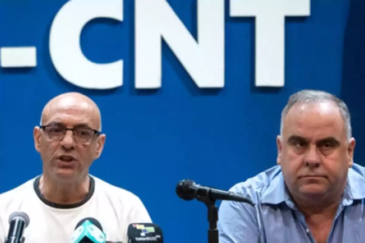 Dos hombres en una conferencia de prensa frente a un fondo azul con las letras "CNT" visibles.