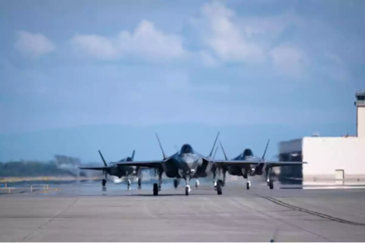 Tres aviones de combate en formación rodando por la pista de un aeropuerto con un cielo azul y nubes al fondo.