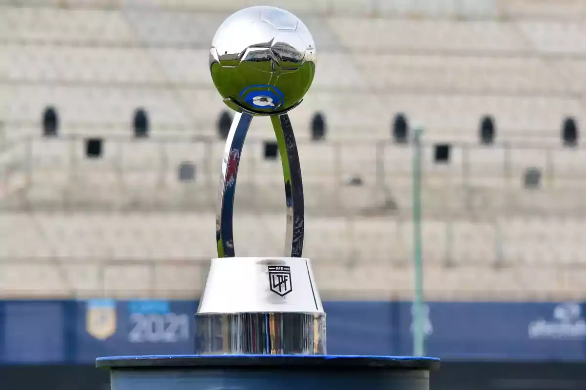 Trofeo de fútbol plateado con forma de balón sobre una base en un estadio vacío.