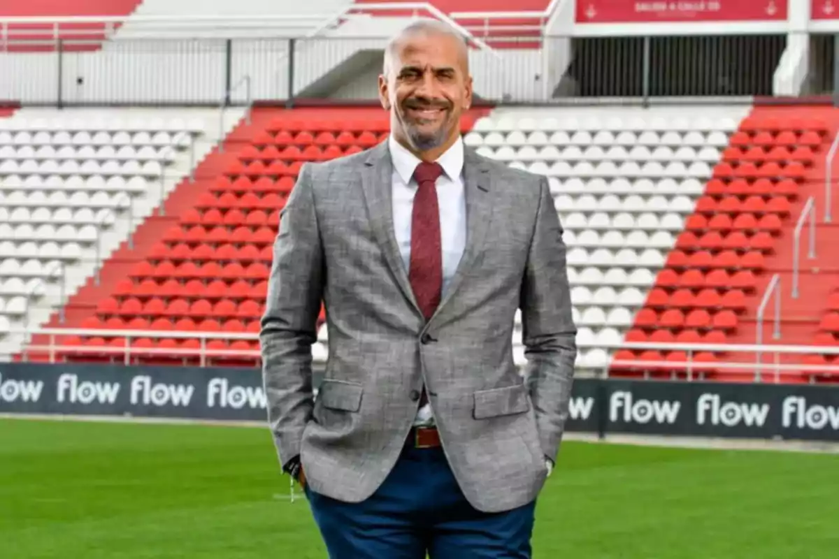 Hombre de pie en un estadio de fútbol con traje gris y corbata roja, sonriendo.