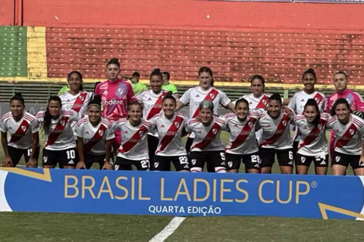 Un equipo de fútbol femenino posa para una foto grupal en el campo, vistiendo uniformes blancos con una banda roja diagonal y detrás de un cartel que dice 