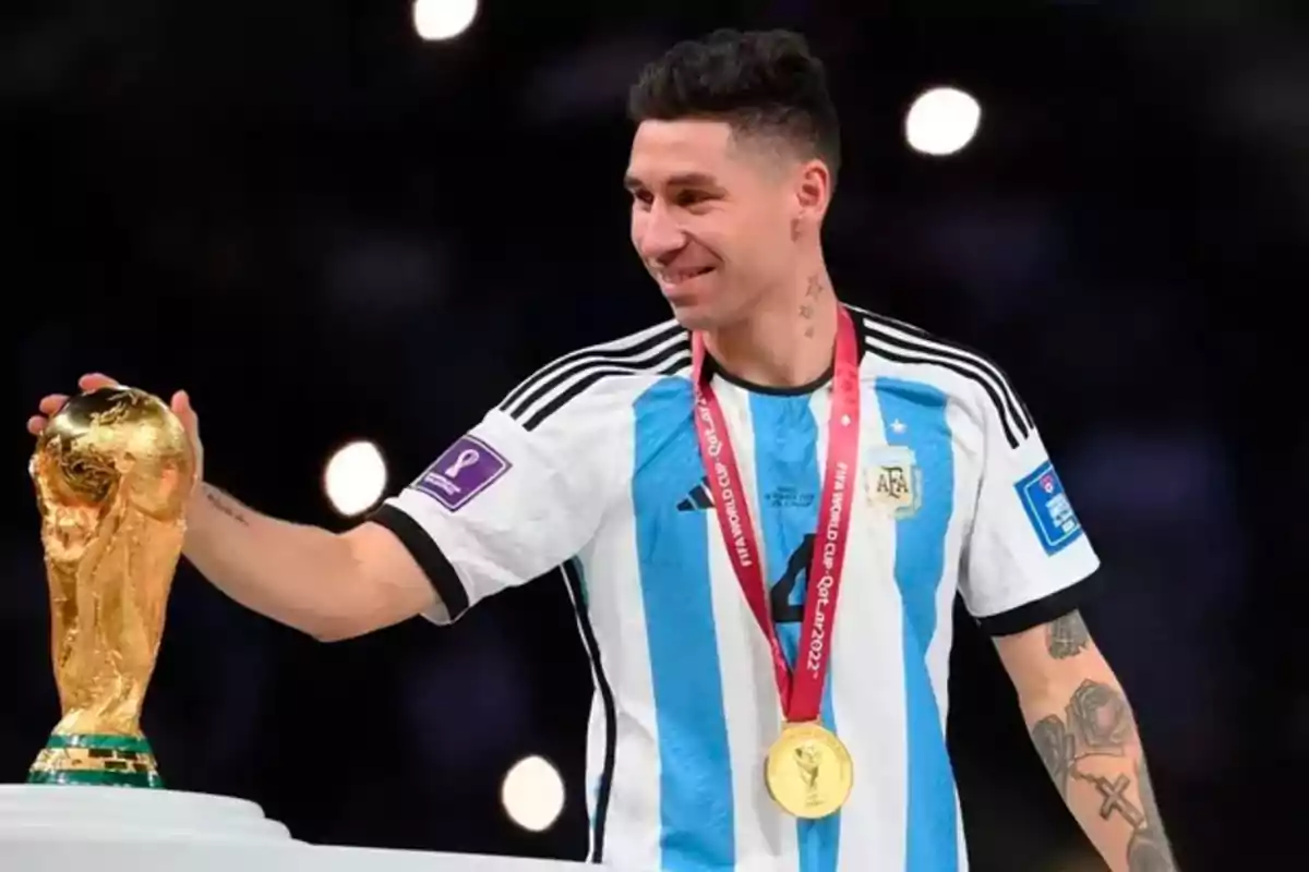 Un jugador de fútbol con la camiseta de Argentina sonríe mientras sostiene el trofeo de la Copa del Mundo y lleva una medalla de oro.