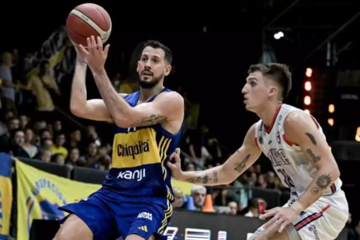 Dos jugadores de baloncesto en acción durante un partido, uno con uniforme azul y amarillo sosteniendo el balón y el otro con uniforme blanco defendiéndolo, con espectadores de fondo.