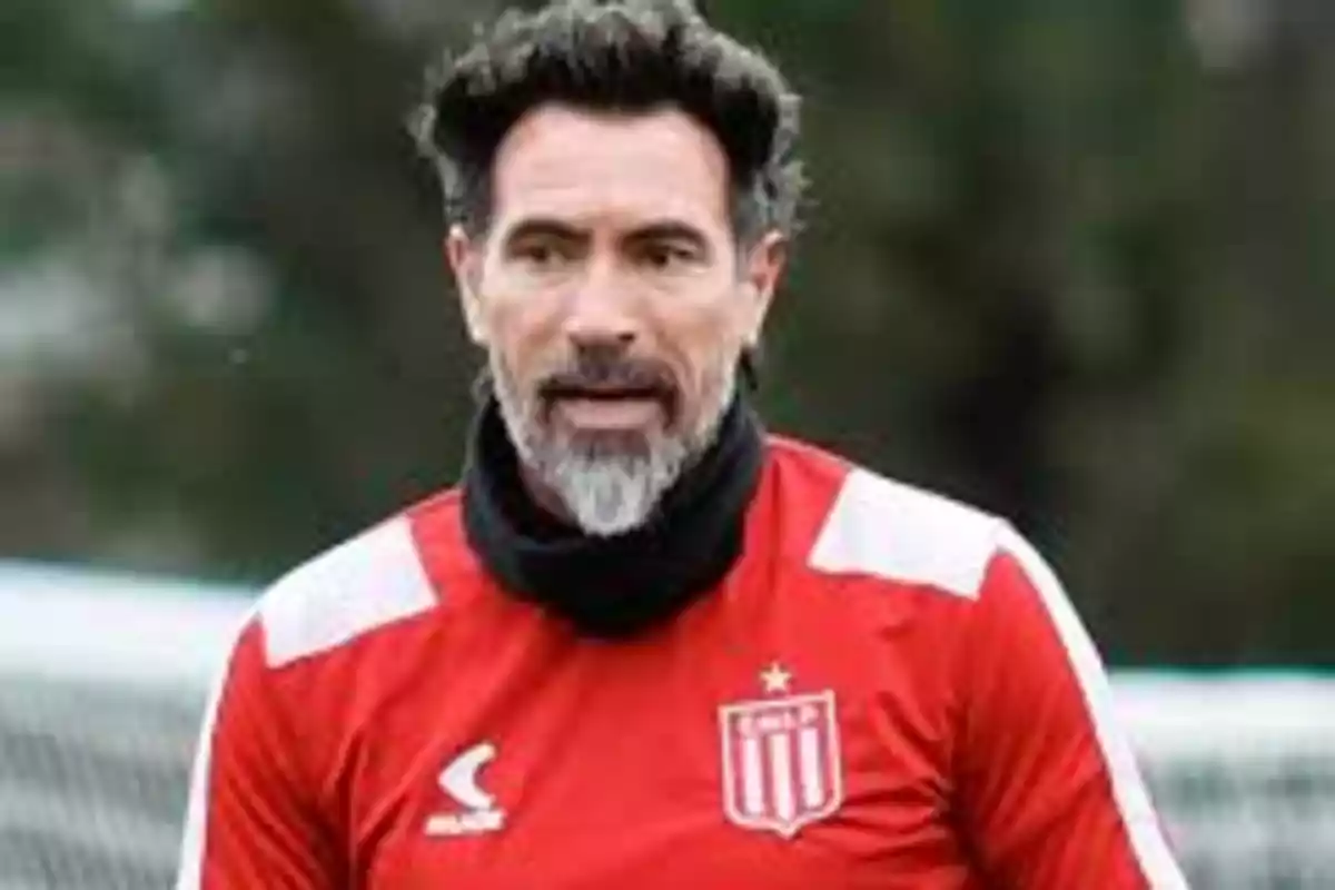 Un hombre con barba y cabello oscuro lleva una camiseta roja con un escudo y está al aire libre.