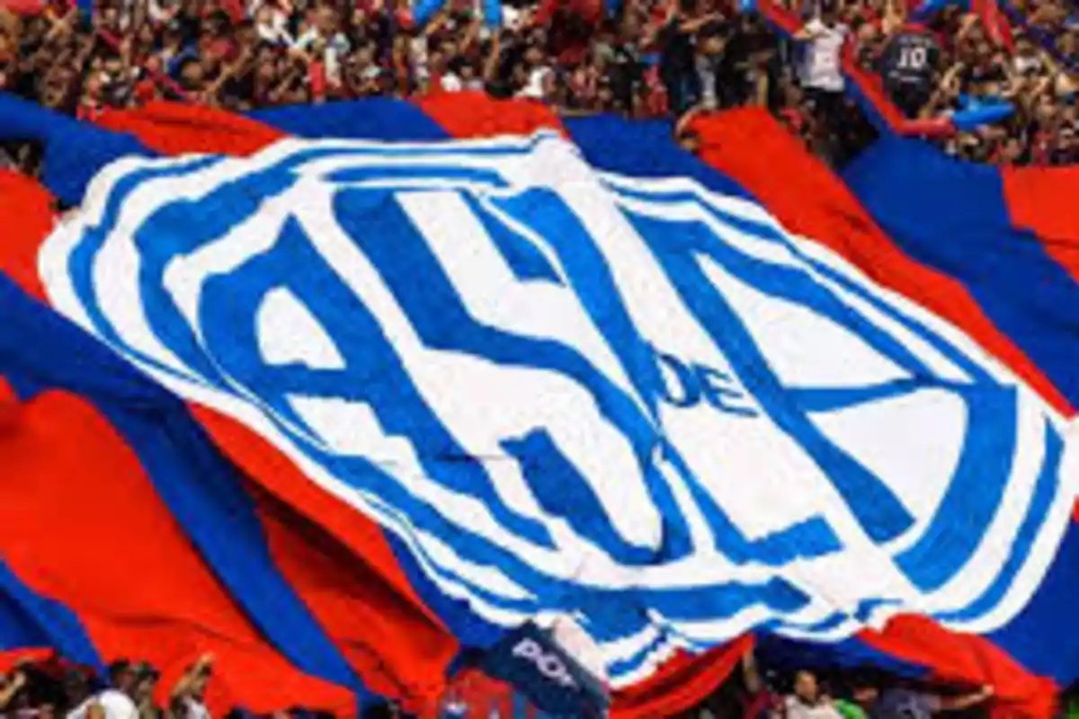 Aficionados sosteniendo una gran bandera azul y roja con un escudo en el centro.