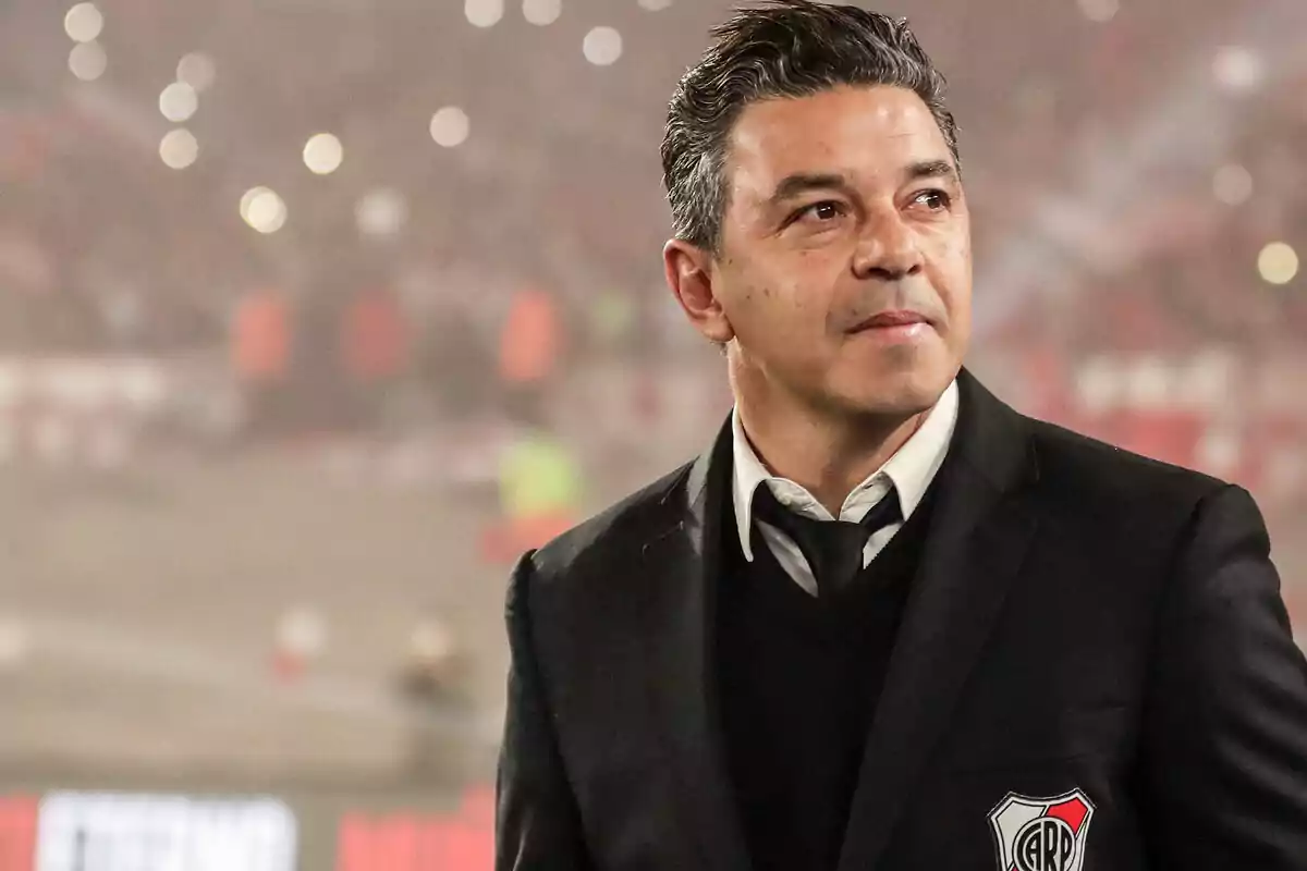 Un hombre con traje oscuro y camisa blanca está de pie en un estadio lleno de luces y espectadores.