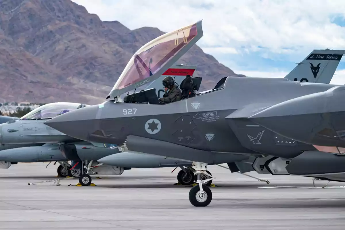 Un avión de combate en una pista con un piloto en la cabina y montañas al fondo.