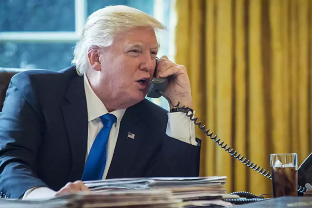 Un hombre de cabello rubio hablando por teléfono en una oficina con cortinas doradas y una bebida sobre el escritorio.