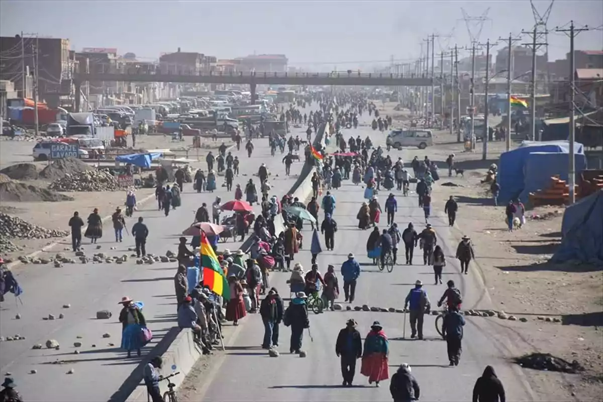 Una multitud de personas caminando por una carretera en una zona urbana con edificios y vehículos al fondo.