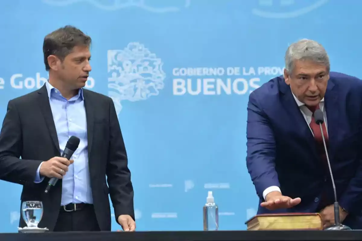 Dos hombres en un evento oficial, uno de ellos sostiene un micrófono mientras el otro realiza un juramento con la mano sobre un libro, con un fondo azul que tiene texto relacionado con el gobierno de la provincia de Buenos Aires.