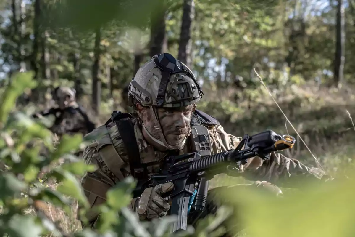 Soldado en uniforme de camuflaje y casco avanzando con cautela en un entorno boscoso.