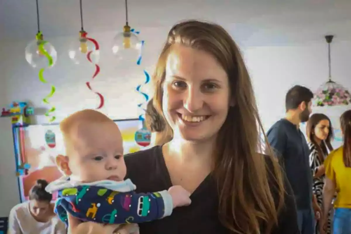 Una mujer sonriente sostiene a un bebé en una fiesta con decoraciones coloridas y personas conversando al fondo.