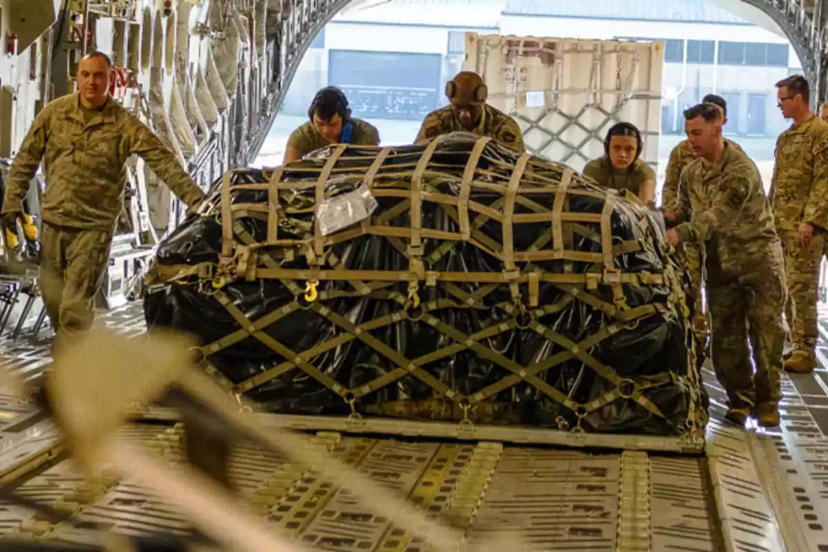 Personal militar cargando un paquete grande y asegurado dentro de un avión de transporte.