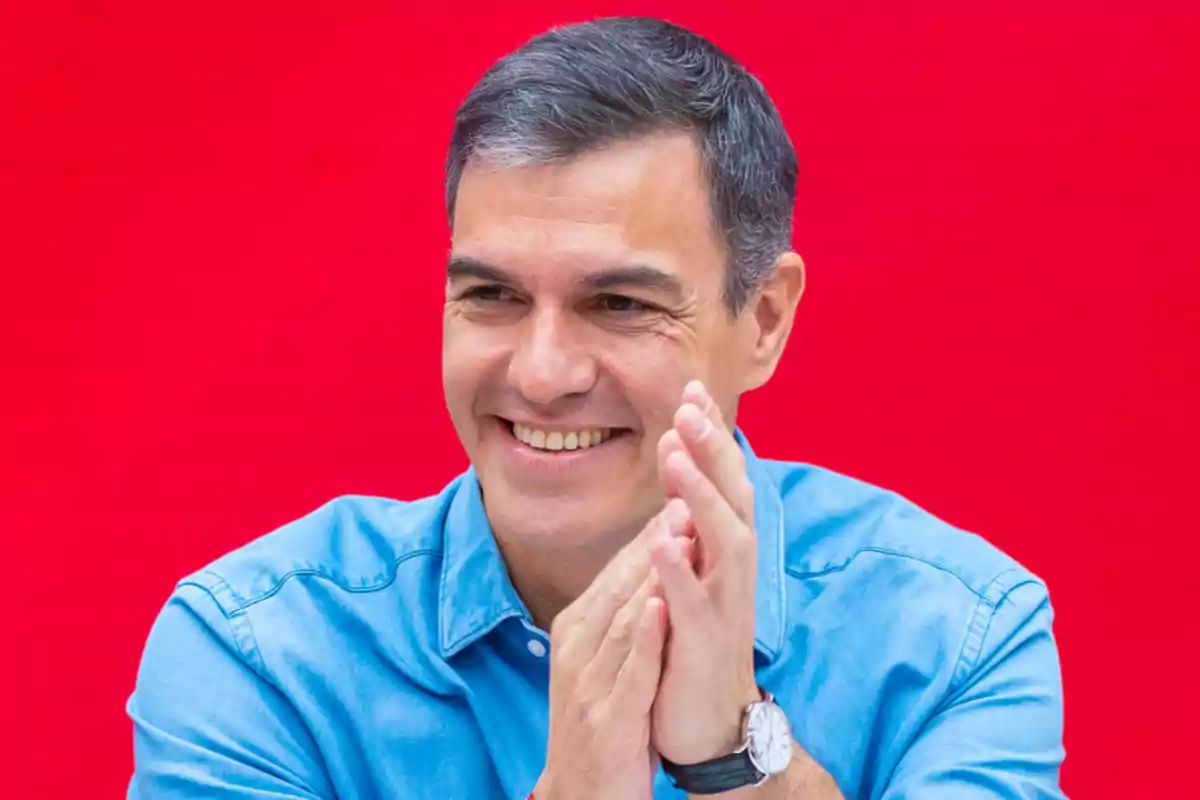 Hombre sonriendo y aplaudiendo con una camisa azul frente a un fondo rojo.