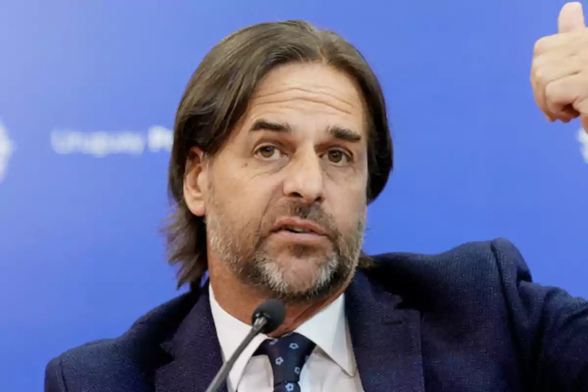 Hombre de cabello castaño y barba hablando en una conferencia de prensa con fondo azul.