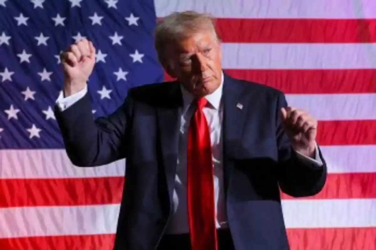 Una persona con traje oscuro y corbata roja gesticulando frente a una bandera de Estados Unidos.