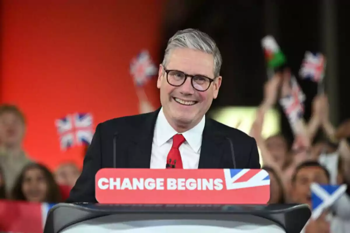 Un hombre sonriente con gafas y traje oscuro habla en un podio con el texto "CHANGE BEGINS" mientras personas al fondo agitan banderas del Reino Unido.