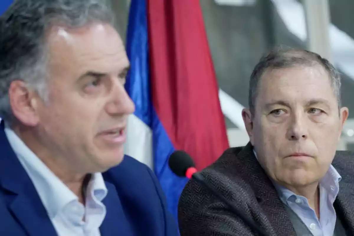 Dos hombres en una conferencia de prensa con una bandera de fondo.