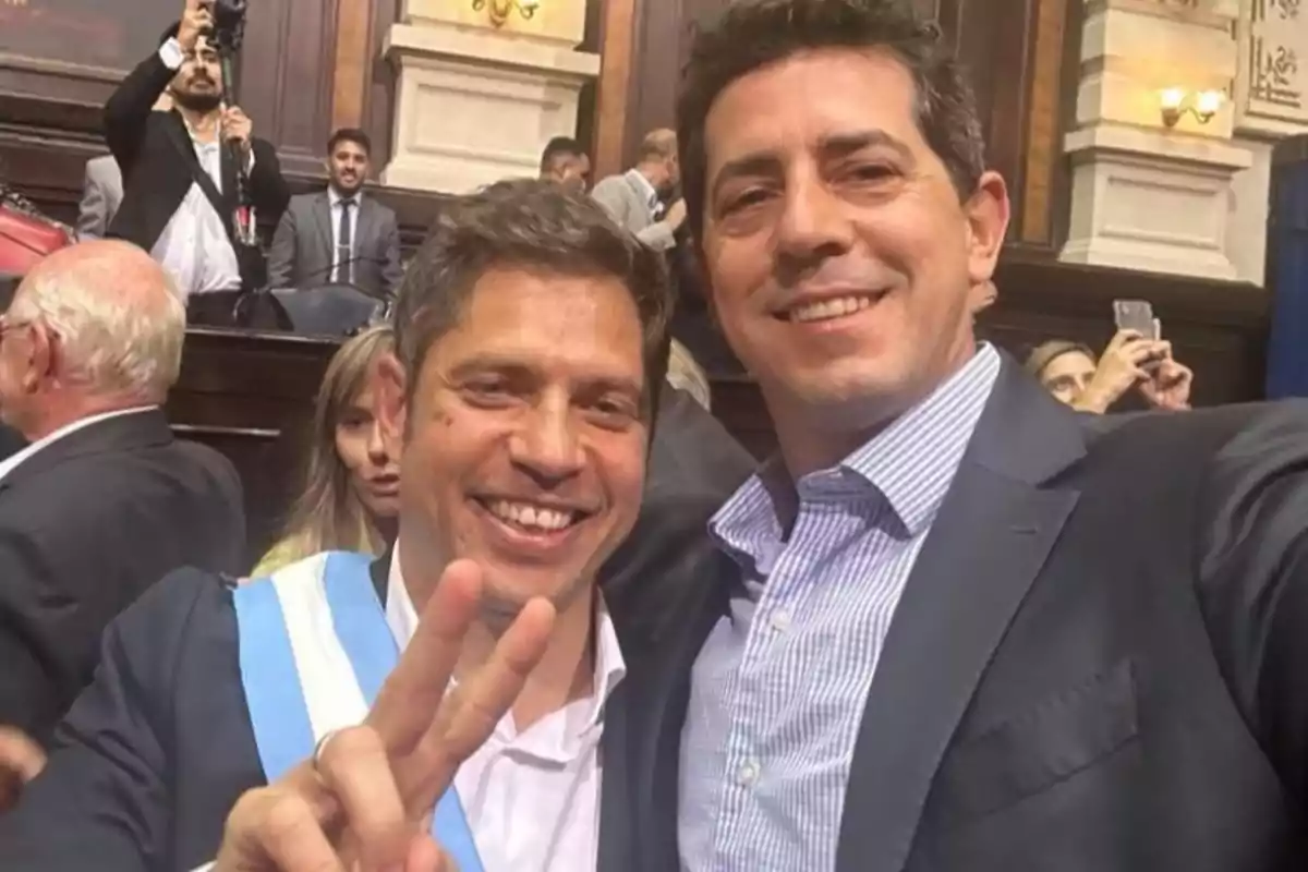 Dos personas sonríen mientras se toman una selfie en un evento formal, una de ellas hace el signo de la paz con los dedos.