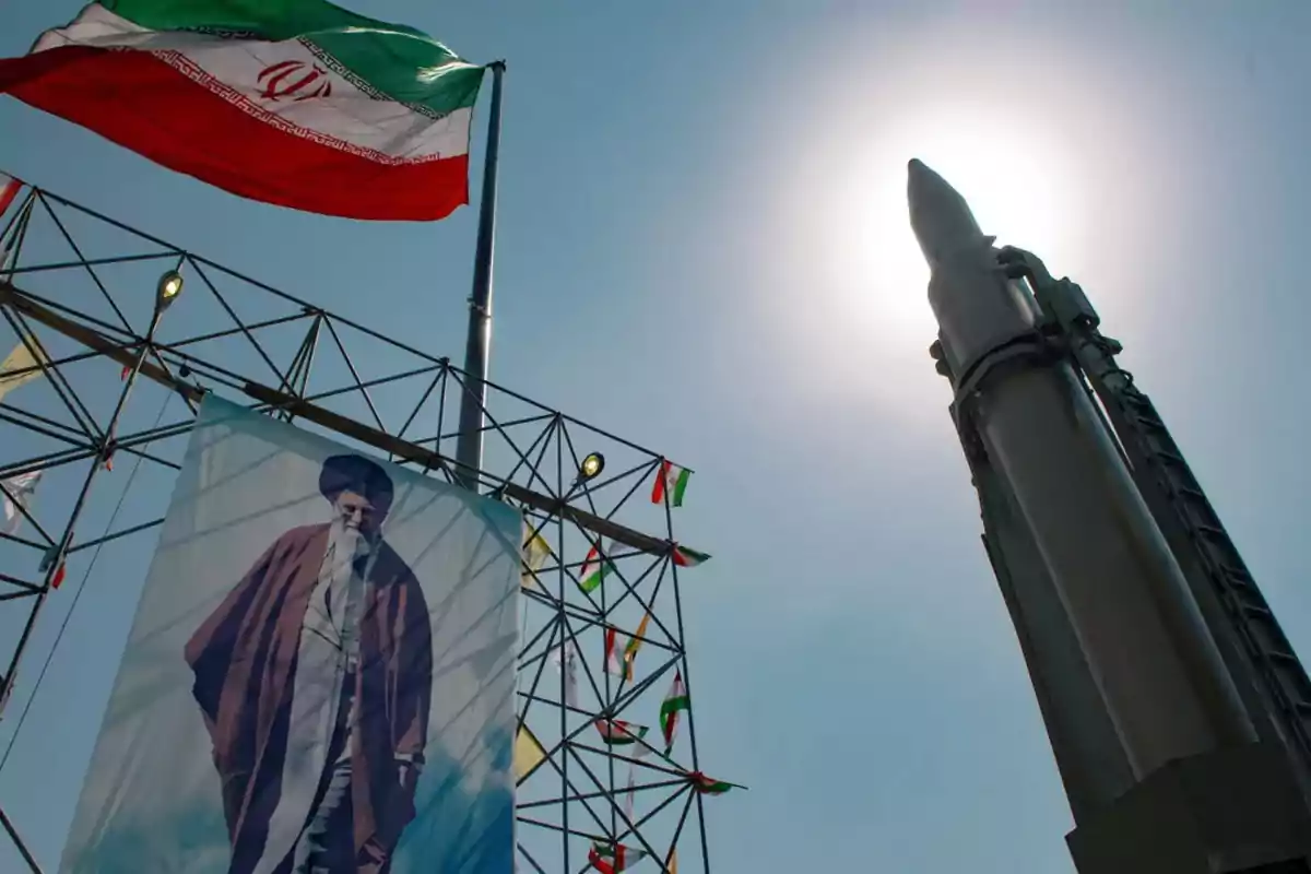 Bandera de Irán ondeando sobre una estructura con un cartel de un líder religioso y un misil en posición vertical iluminado por el sol.