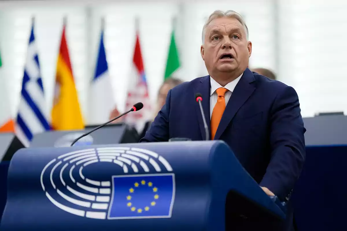 Un hombre de traje azul y corbata naranja habla en un podio con el emblema de la Unión Europea, con banderas de varios países al fondo.