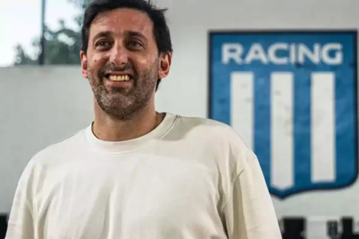 Hombre sonriendo con una camiseta blanca frente a un escudo de Racing Club.