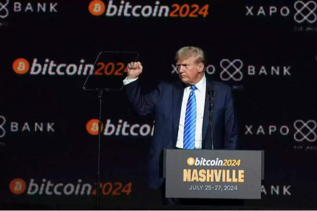 Un hombre de traje y corbata azul está en un escenario con un fondo que muestra logotipos de Bitcoin 2024 y Xapo Bank mientras levanta el puño junto a un podio que indica un evento en Nashville del 25 al 27 de julio de 2024.