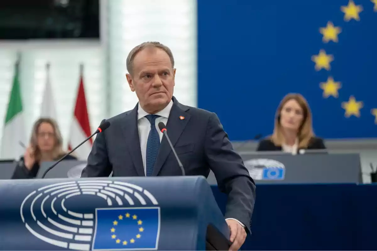 Un hombre de traje habla en un podio con el emblema de la Unión Europea en un entorno de conferencia con banderas y personas al fondo.