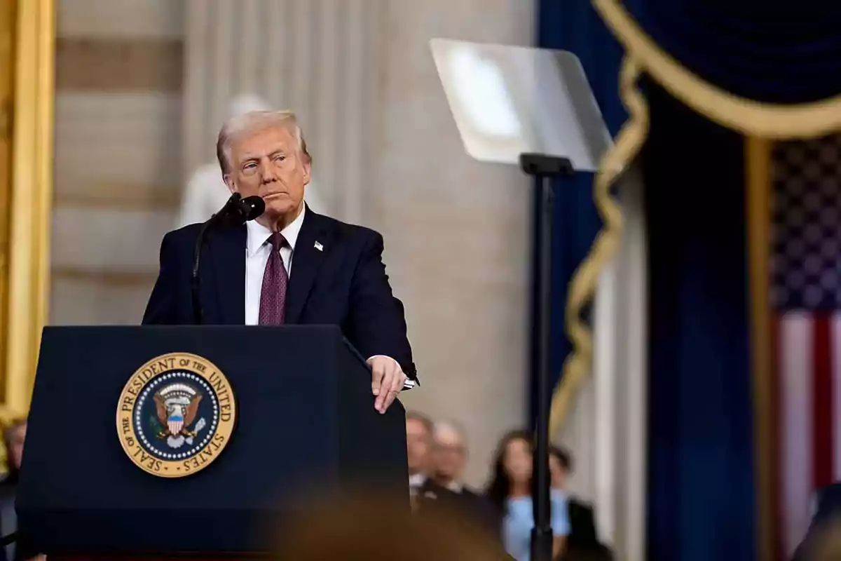 Un hombre de traje oscuro y corbata roja está de pie detrás de un podio con un sello presidencial, hablando frente a un micrófono en un entorno formal con una bandera estadounidense al fondo.