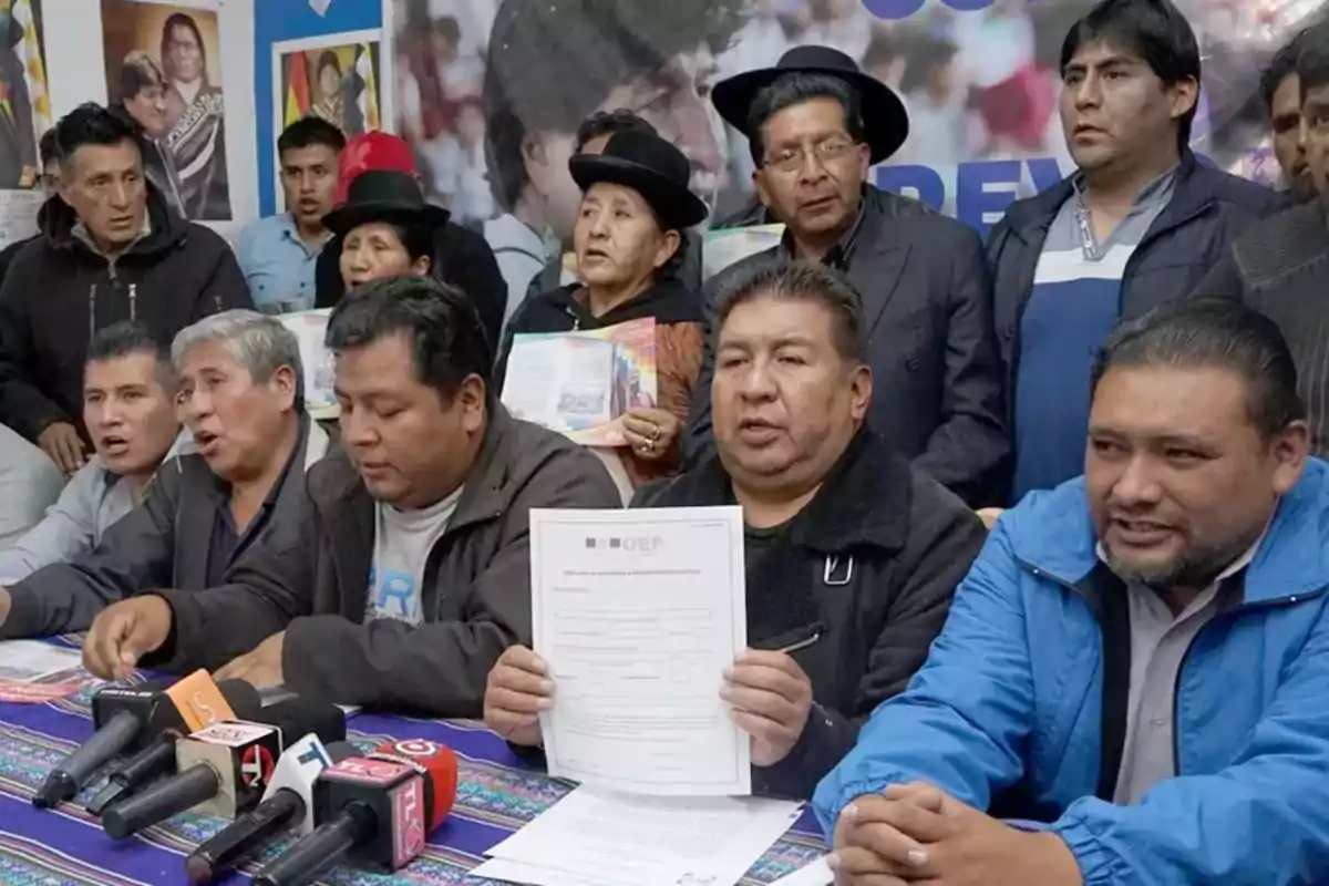 A group of people at a press conference with microphones and documents on a table.