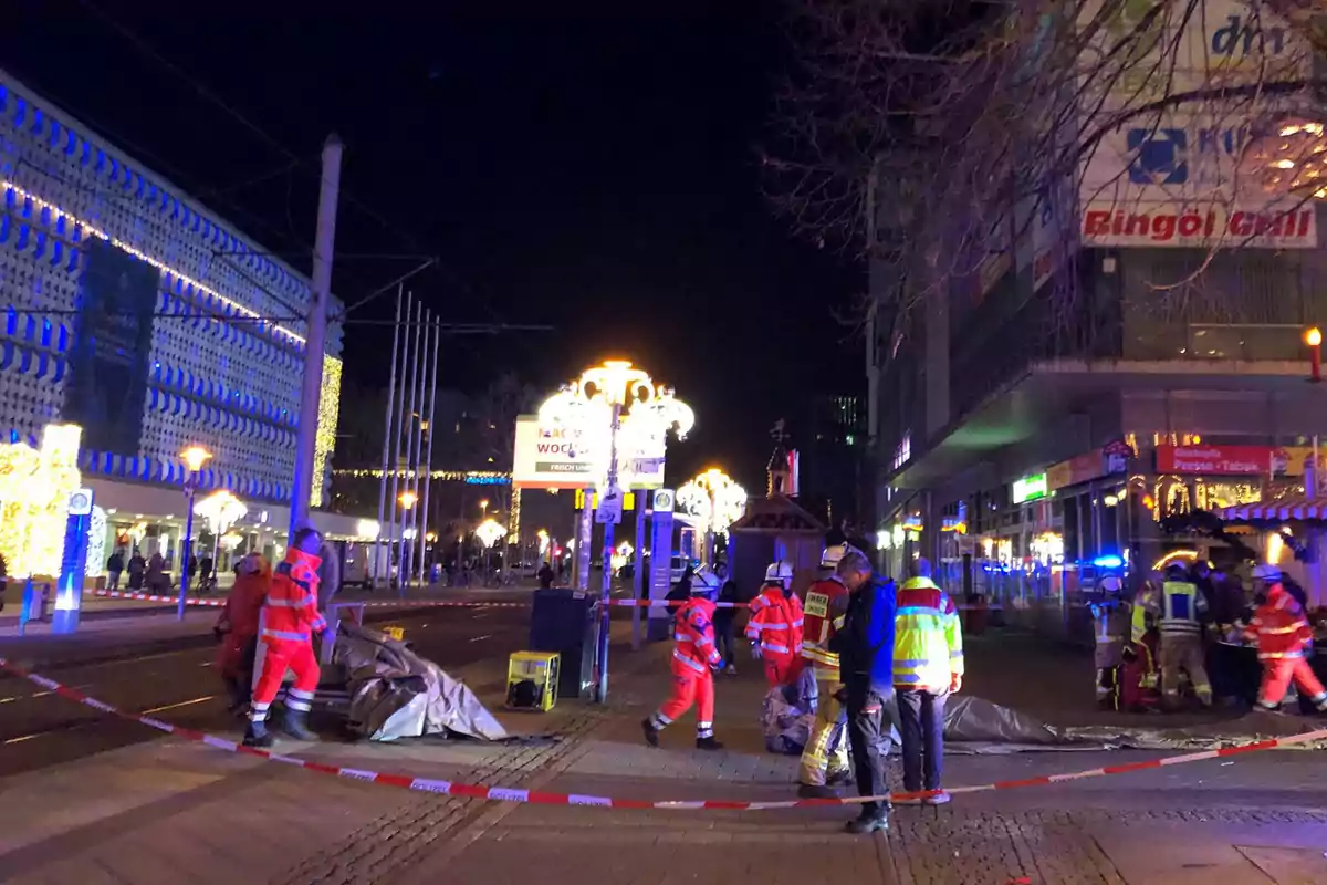 Un grupo de personas con chalecos reflectantes y uniformes de emergencia se encuentra en una calle acordonada por la noche, con luces brillantes y edificios iluminados al fondo.