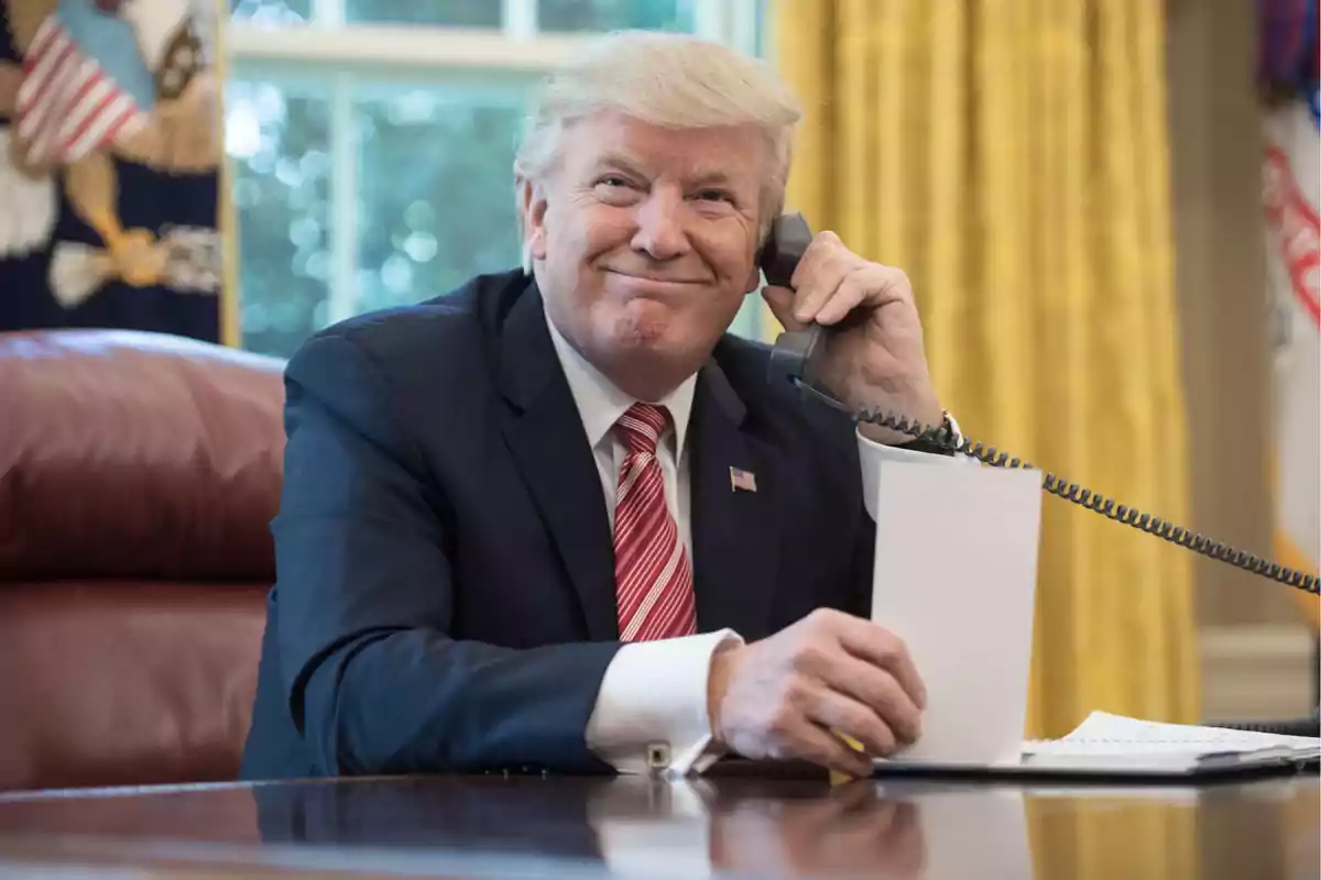 Un hombre de traje oscuro y corbata roja está sentado en un escritorio hablando por teléfono con una expresión sonriente.