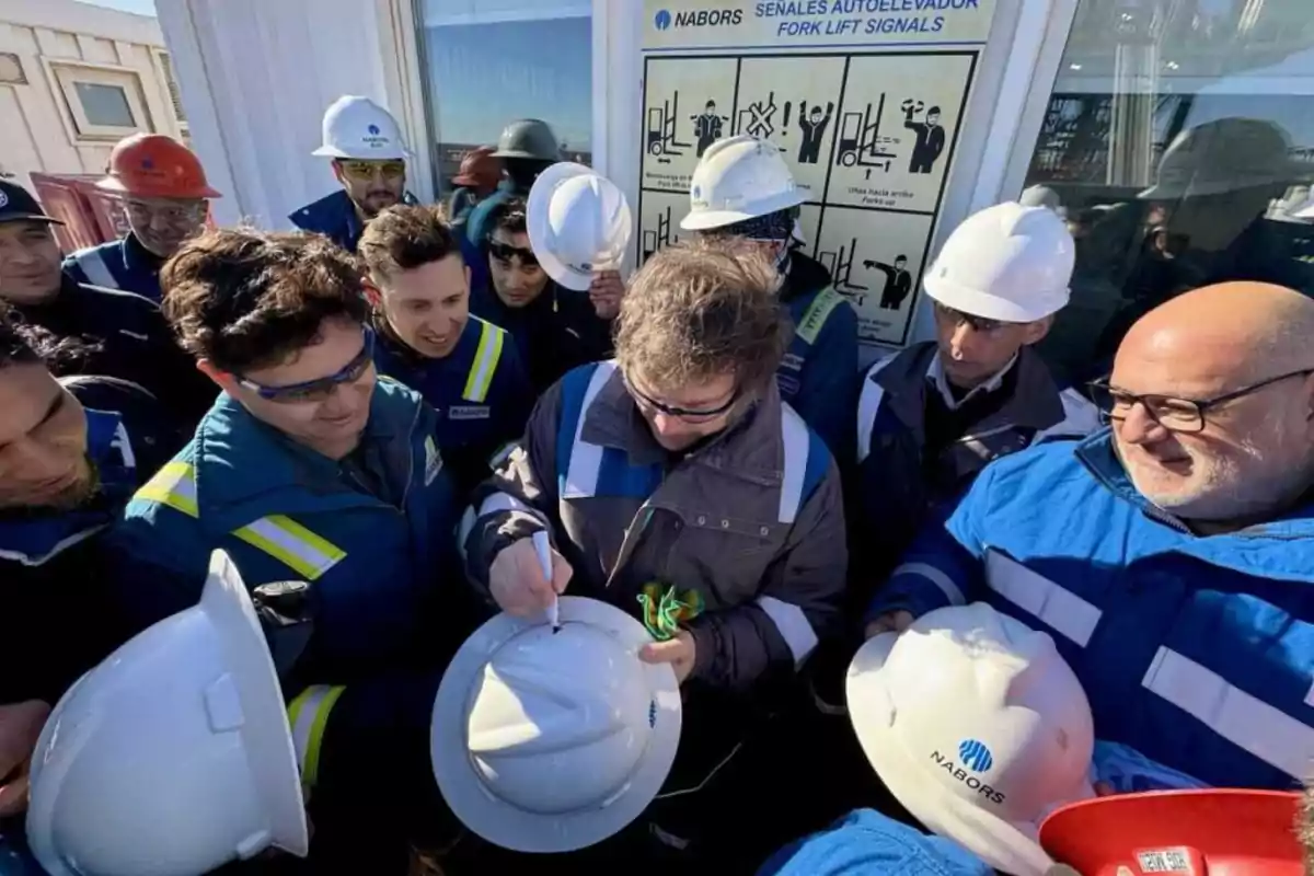 Un grupo de trabajadores con cascos de seguridad rodea a una persona que está firmando un casco blanco, con señales de seguridad para montacargas visibles en el fondo.