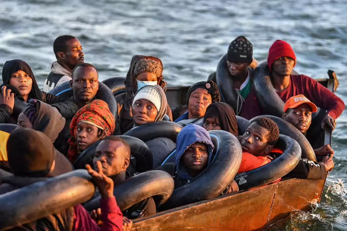 Un grupo de personas en un bote sobre el agua, algunas llevan gorros y bufandas, y están rodeadas de neumáticos inflables.