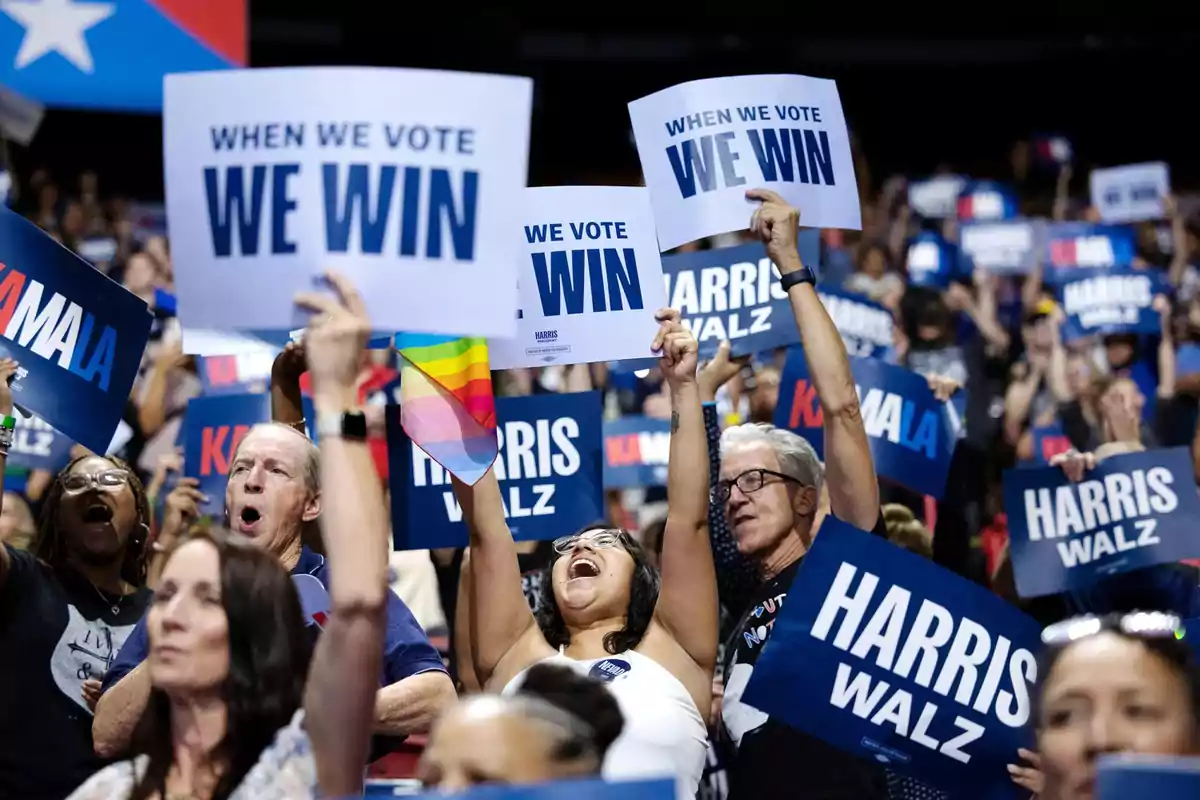 Personas en un mitin político sosteniendo carteles que dicen "WHEN WE VOTE WE WIN" y "HARRIS WALZ" mientras muestran entusiasmo y apoyo.