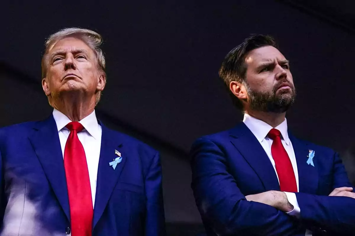 Two men standing in dark suits and red ties, looking forward with serious expressions.