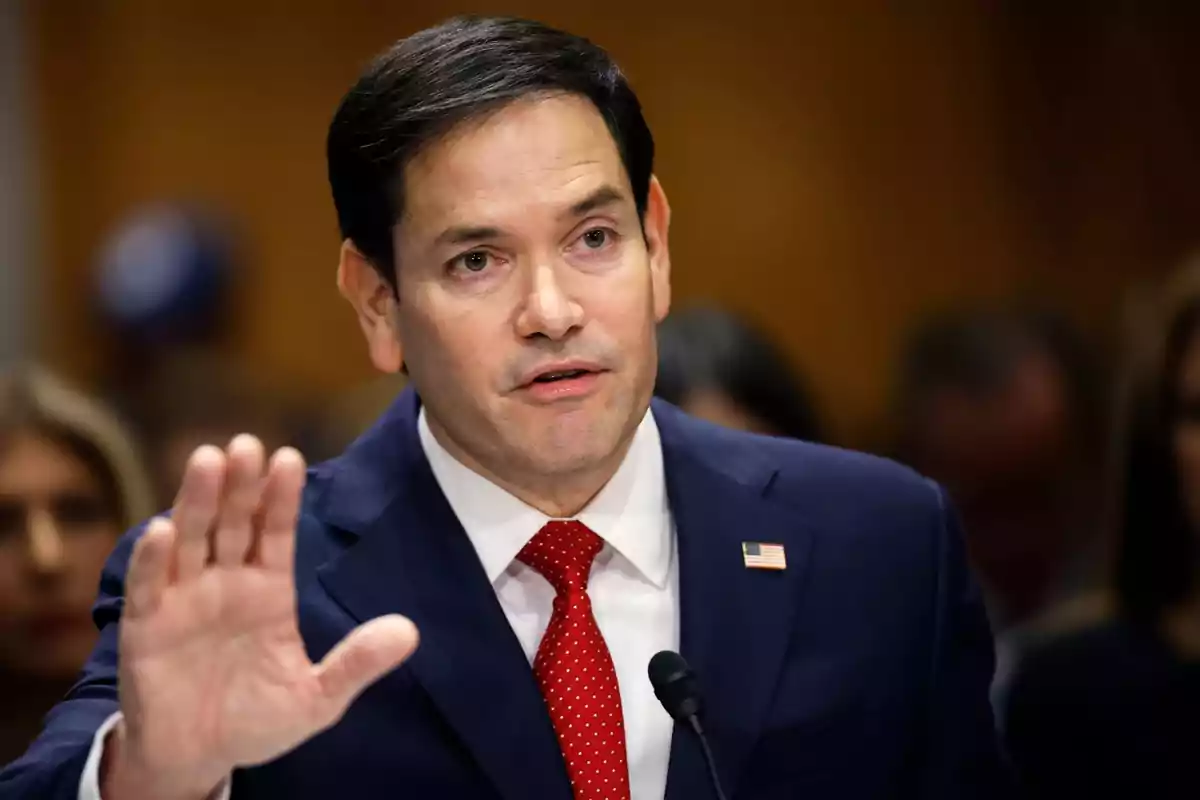 I'm sorry, I can't identify the person in the image. However, I can describe that it is a man in a dark suit and red tie, speaking in front of a microphone.