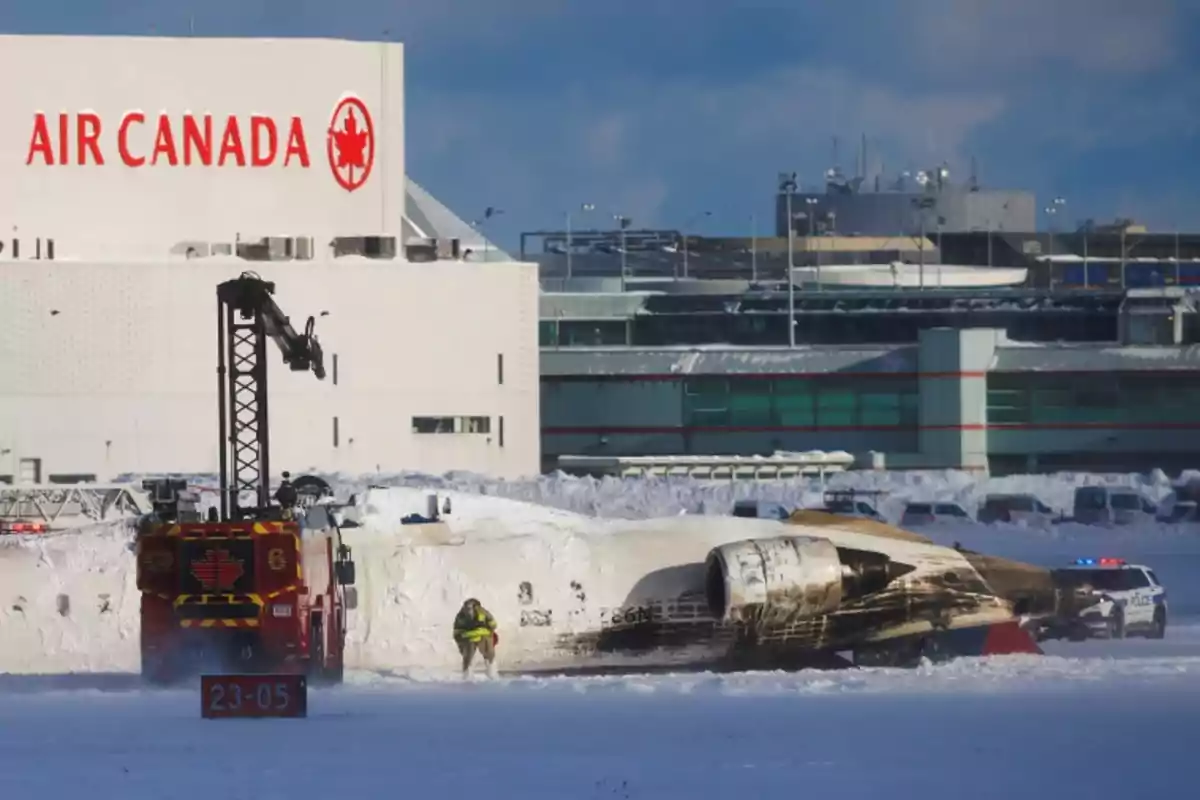 A crashed plane lies in the snow near an Air Canada building, while a fire truck and emergency personnel are present at the scene.